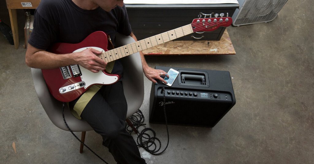 A man with an electric guitar, using a smart phone with his practice guitar amp